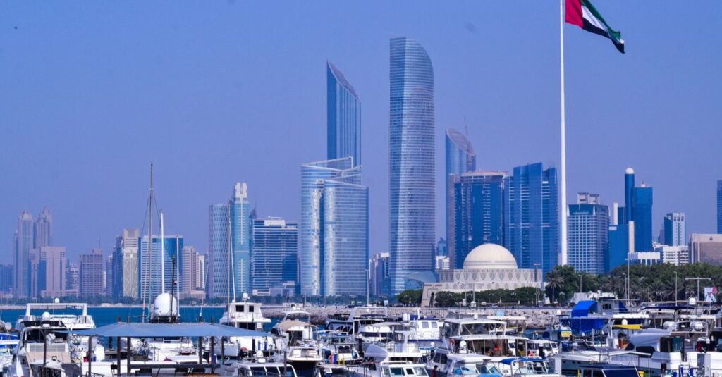 A scenic view of Abu Dhabi marina with modern skyscrapers and a flag in the foreground.